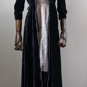 K&ouml;nnte beinhalten: Eine lange schwarze Samt-Robe mit einem Rei&szlig;verschluss an der Vorderseite. Die Robe ist offen und zeigt darunter ein wei&szlig;es Spitzenkleid.