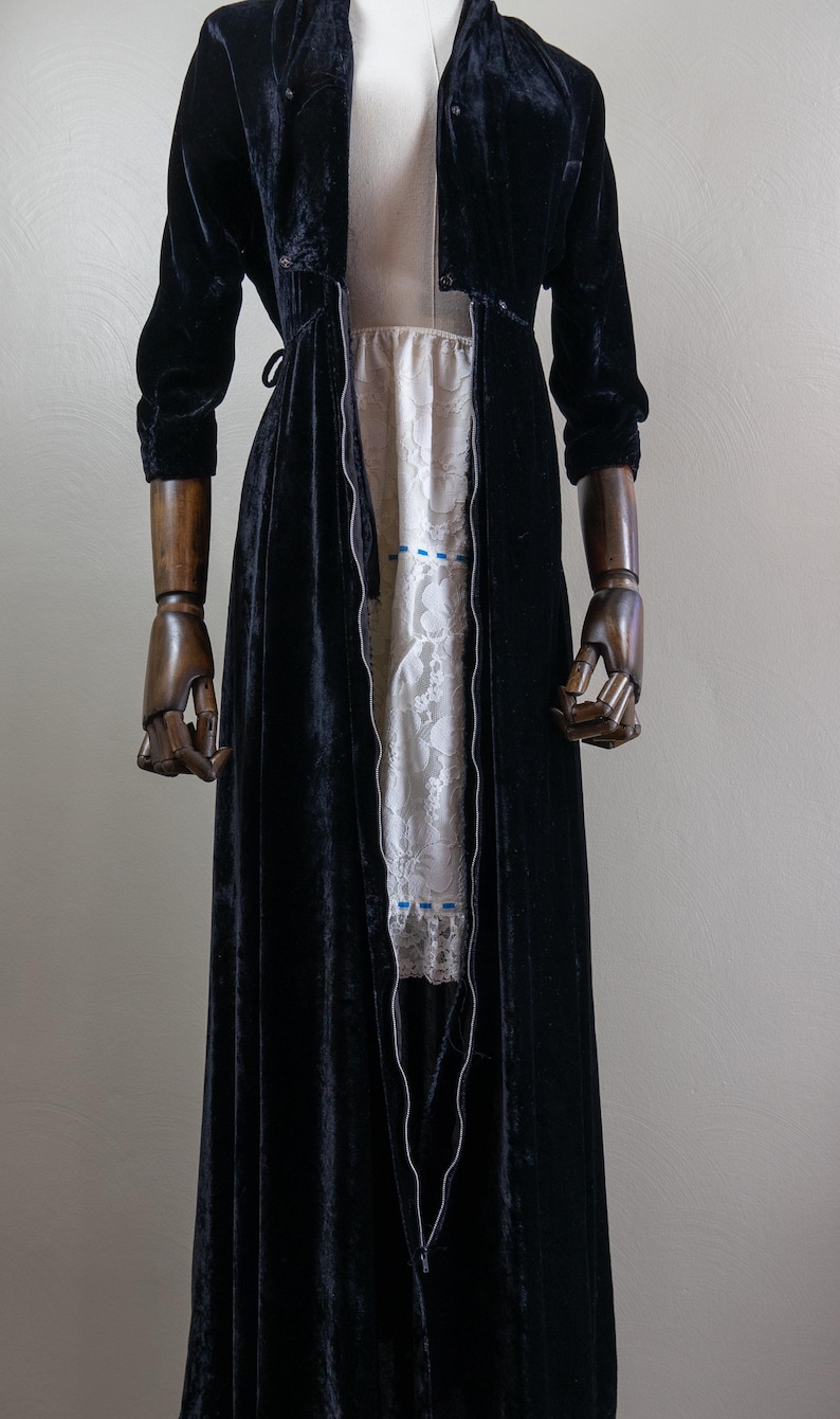 K&ouml;nnte beinhalten: Eine lange schwarze Samt-Robe mit einem Rei&szlig;verschluss an der Vorderseite. Die Robe ist offen und zeigt darunter ein wei&szlig;es Spitzenkleid.