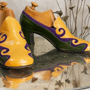May include: A pair of vintage women's shoes with wooden shoe forms. The shoes feature a yellow toe and vamp, green sides, and purple accents. The heels are black. The shoes are displayed on a reflective surface.