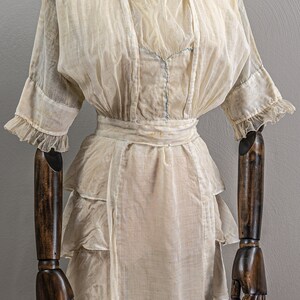 May include: A vintage white sheer dress with a ruffled neckline and sleeves. The dress has a fitted bodice and a flowing skirt with multiple layers of ruffles. The dress is displayed on a wooden dress form.