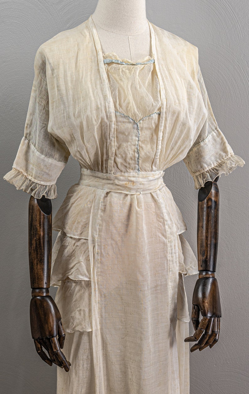 May include: A vintage white sheer dress with a ruffled neckline and sleeves. The dress has a fitted bodice and a flowing skirt with multiple layers of ruffles. The dress is displayed on a wooden dress form.