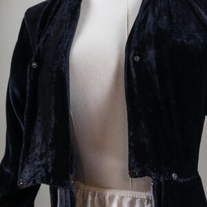 K&ouml;nnte beinhalten: Eine schwarze Samtjacke mit Rei&szlig;verschluss und Knopfdetails. Die Jacke hat eine lange, flie&szlig;ende Silhouette und eine lockere Passform.