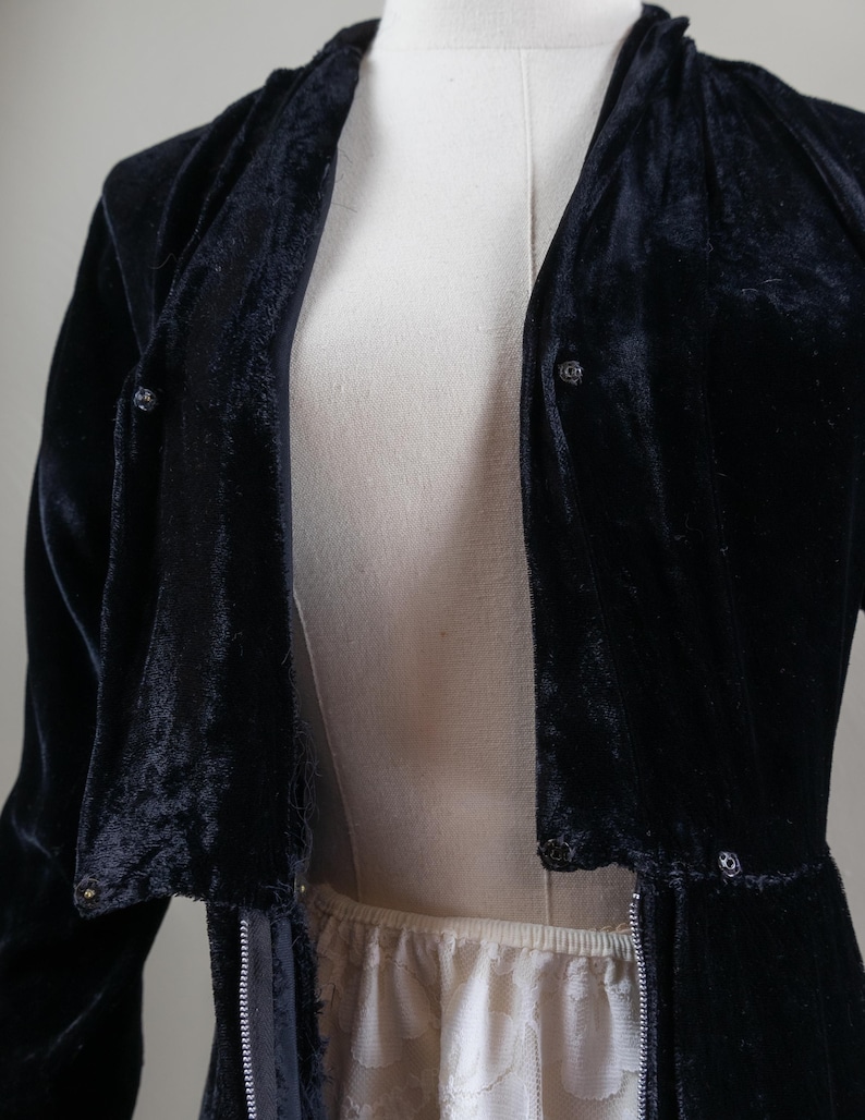 K&ouml;nnte beinhalten: Eine schwarze Samtjacke mit Rei&szlig;verschluss und Knopfdetails. Die Jacke hat eine lange, flie&szlig;ende Silhouette und eine lockere Passform.