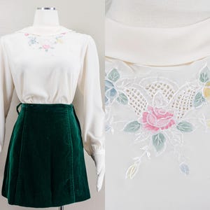 May include: A vintage cream-colored blouse with long sleeves and a green velvet skirt. The blouse features a floral embroidered collar with pink and yellow roses. The skirt is a deep forest green, with a high waist and a slight A-line shape.