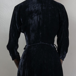 K&ouml;nnte beinhalten: Ein schwarzes Samtkleid mit langem Rock und langen &Auml;rmeln. Das Kleid hat einen Wickelstil und wird in der Taille gebunden.