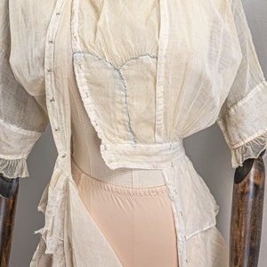 May include: A white sheer dress with a lace overlay and a blue ribbon detail. The dress is on a mannequin and has a ruffled hem.