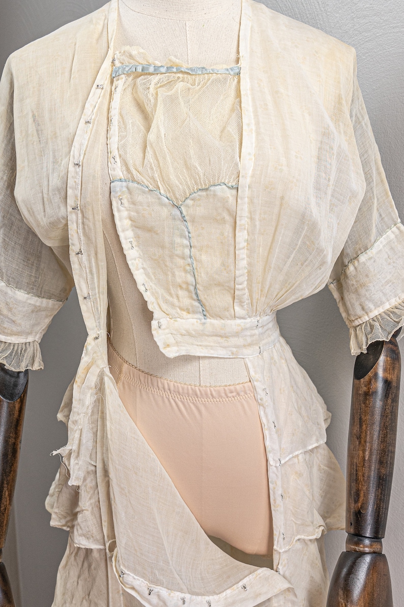 May include: A white sheer dress with a lace overlay and a blue ribbon detail. The dress is on a mannequin and has a ruffled hem.