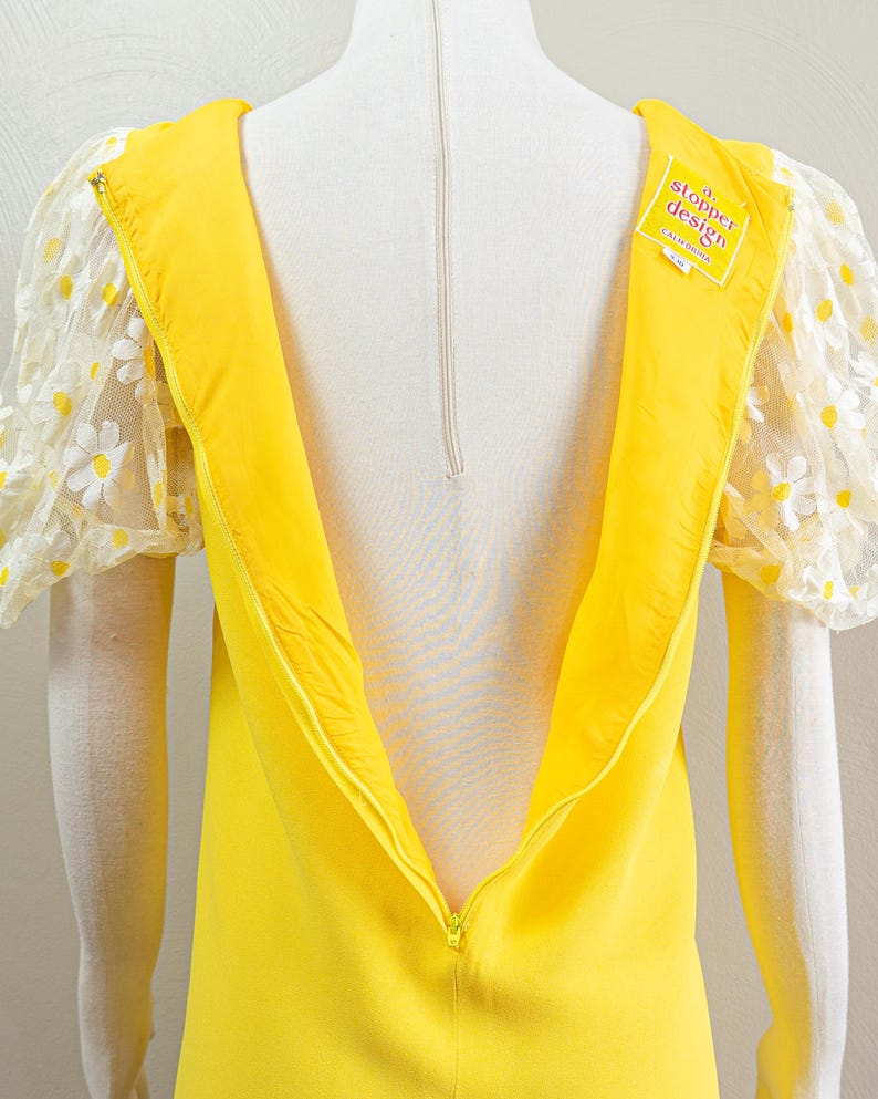 May include: A bright yellow dress with a zipper down the back. The dress features sheer, puffy sleeves adorned with white daisy appliqu&eacute;s. A tag on the dress reads "a. stopper design California".