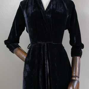 K&ouml;nnte beinhalten: Ein schwarzes Samtkleid mit V-Ausschnitt und langen &Auml;rmeln. Das Kleid hat einen G&uuml;rtel an der Taille und einen Rei&szlig;verschluss vorne.