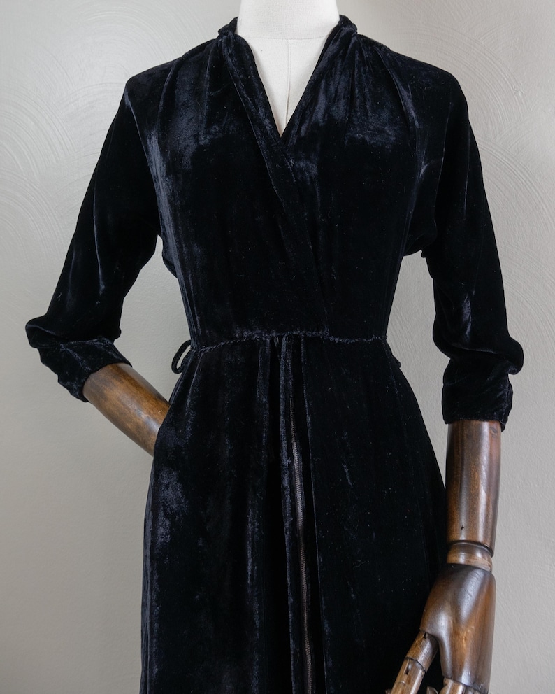 K&ouml;nnte beinhalten: Ein schwarzes Samtkleid mit V-Ausschnitt und langen &Auml;rmeln. Das Kleid hat einen G&uuml;rtel an der Taille und einen Rei&szlig;verschluss vorne.
