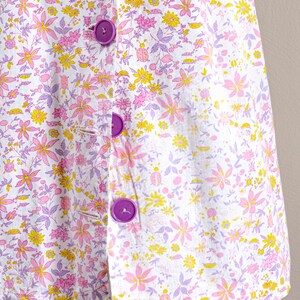 Puede incluir: Prenda con estampado floral con fondo blanco adornado con flores rosas, amarillas y moradas. Cuenta con grandes botones morados. El tejido tiene una est&eacute;tica delicada y femenina.