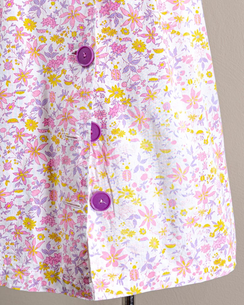 Puede incluir: Prenda con estampado floral con fondo blanco adornado con flores rosas, amarillas y moradas. Cuenta con grandes botones morados. El tejido tiene una est&eacute;tica delicada y femenina.