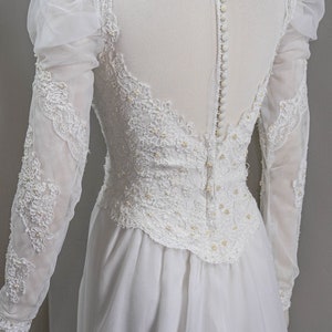 May include: A white wedding dress with long sleeves and a lace bodice. The dress has a high neckline and a button-up back.