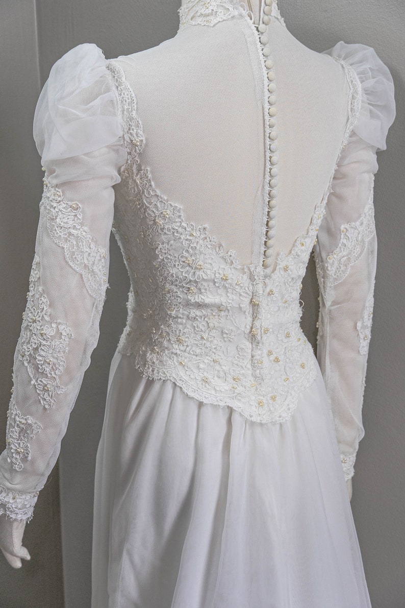 May include: A white wedding dress with long sleeves and a lace bodice. The dress has a high neckline and a button-up back.