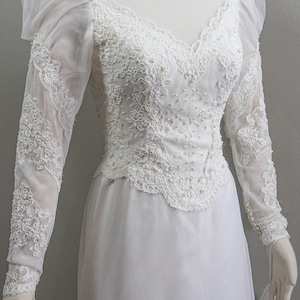 May include: A white wedding dress with long sleeves and a high neckline. The dress is made of lace and features a fitted bodice and a flowing skirt.