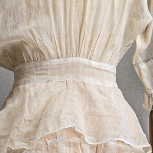 May include: A white sheer blouse with a gathered waist and a ruffled hem. The blouse has a delicate floral pattern.