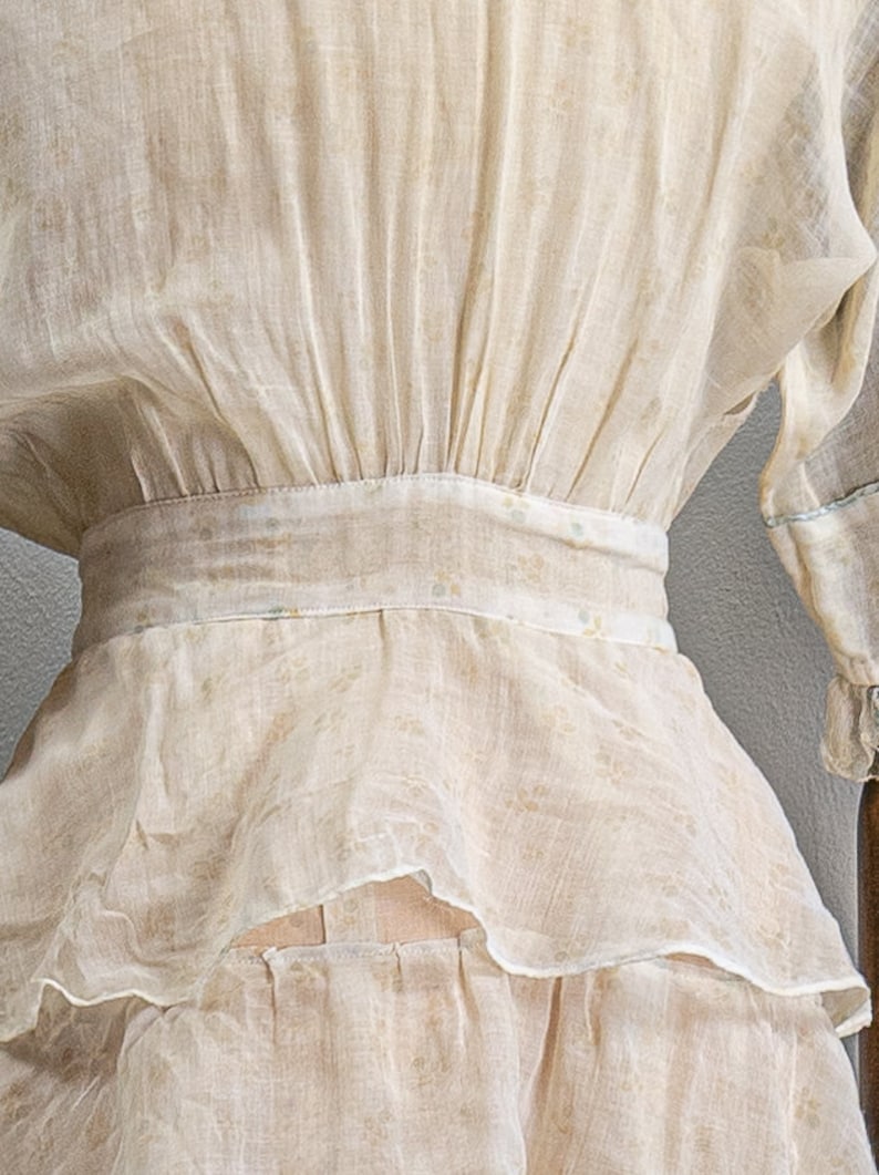 May include: A white sheer blouse with a gathered waist and a ruffled hem. The blouse has a delicate floral pattern.