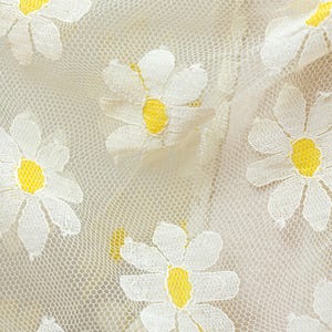 May include: Close-up of a cream-colored lace fabric with embroidered white daisies featuring yellow centers. The delicate floral pattern is repeated across the fabric, creating a textured and visually appealing design.