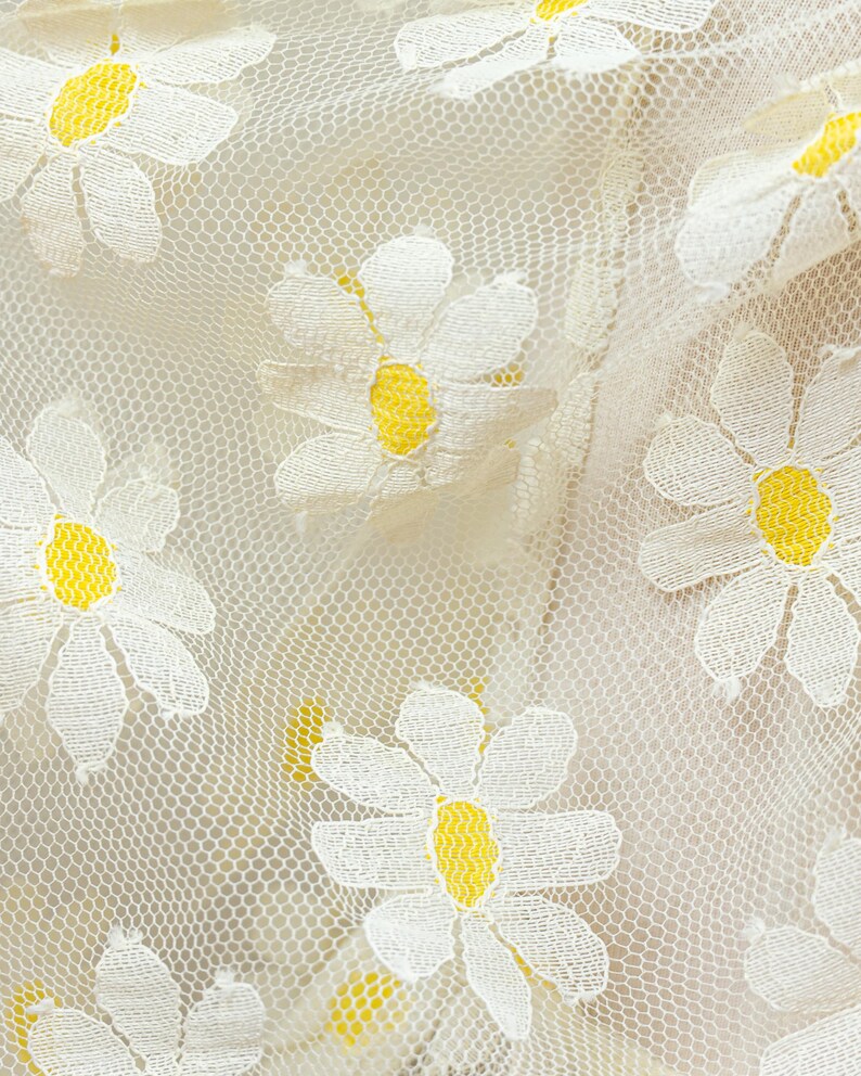 May include: Close-up of a cream-colored lace fabric with embroidered white daisies featuring yellow centers. The delicate floral pattern is repeated across the fabric, creating a textured and visually appealing design.