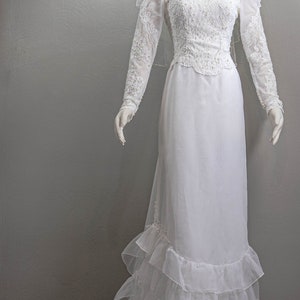 May include: A white wedding dress with long sleeves, a lace bodice, and a ruffled skirt. The dress is on a mannequin and has a veil.