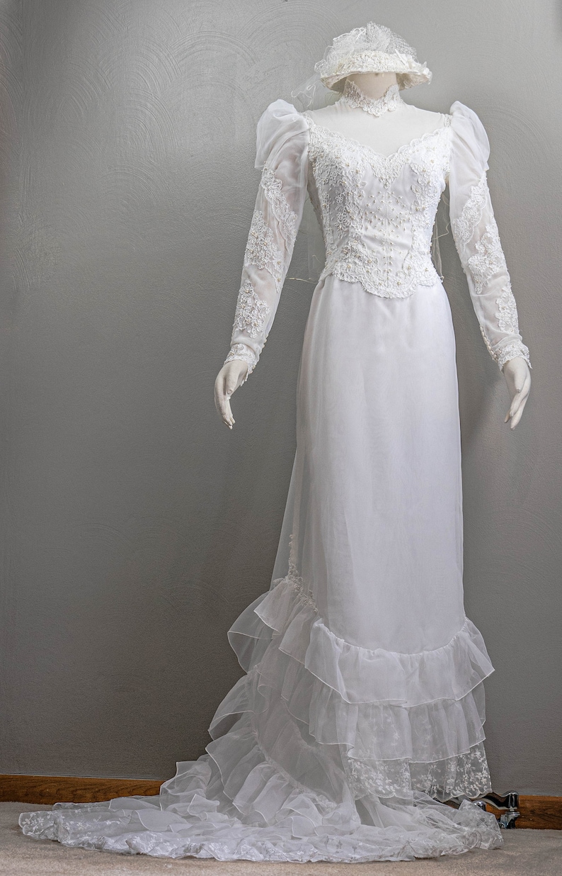 May include: A white wedding dress with long sleeves, a lace bodice, and a ruffled skirt. The dress is on a mannequin and has a veil.