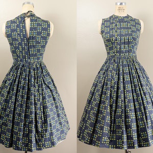 May include: A vintage-style sleeveless dress in a dark blue fabric with a repeating pattern of green and light blue squares. The dress features a fitted bodice, a high neckline, and a flared skirt. The dress is displayed on a mannequin.