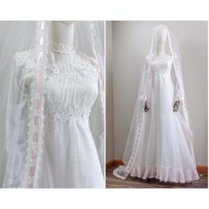 Puede incluir: Un vestido de novia vintage blanco con mangas largas y un corpiño de encaje. El vestido tiene un ribete de cinta rosa y un velo transparente.