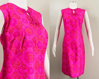 May include: A sleeveless, knee-length dress in a vibrant fuchsia hue, adorned with a repeating floral pattern in shades of orange and pink. The dress features a keyhole neckline with decorative detailing.