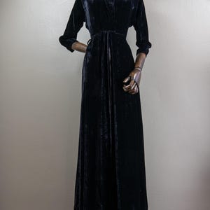 K&ouml;nnte beinhalten: Ein langes schwarzes Samtkleid mit V-Ausschnitt und langen &Auml;rmeln. Das Kleid hat ein tailliertes Oberteil und einen flie&szlig;enden Rock.