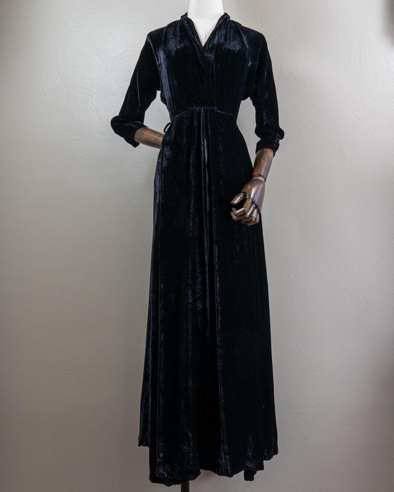 K&ouml;nnte beinhalten: Ein langes schwarzes Samtkleid mit V-Ausschnitt und langen &Auml;rmeln. Das Kleid hat ein tailliertes Oberteil und einen flie&szlig;enden Rock.