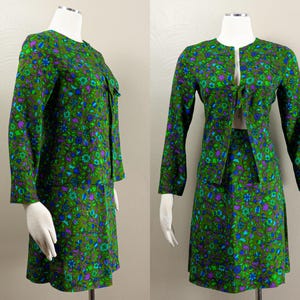 Puede incluir: Un conjunto de dos piezas compuesto por una chaqueta de manga larga y una falda a juego, ambas con un estampado floral en tonos verde oliva, azul brillante y morado. La chaqueta presenta un cuello redondo y un cierre de lazo frontal.