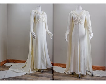 May include: A white floor-length gown with a long train and a hooded cape. The gown has a fitted bodice and a flowing skirt.