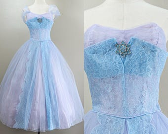 May include: A vintage light blue and lavender formal dress. The bodice features a lace overlay and a jeweled brooch. The skirt is full, with a tulle overlay and lace accents. The dress is displayed on a mannequin.