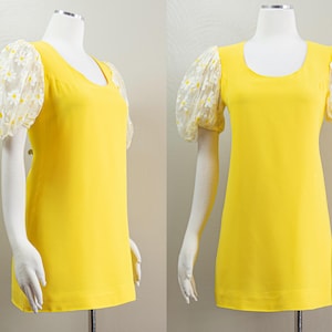 May include: A bright yellow dress with a scoop neckline and short, sheer sleeves adorned with white daisy embroidery. The dress is displayed on a mannequin, showcasing its A-line silhouette.