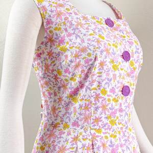 Puede incluir: Vestido sin mangas con fondo blanco y estampado floral en rosa, amarillo y morado. El vestido presenta un escote cuadrado, grandes botones morados y un corpi&ntilde;o entallado. El vestido se exhibe en un maniqu&iacute;.