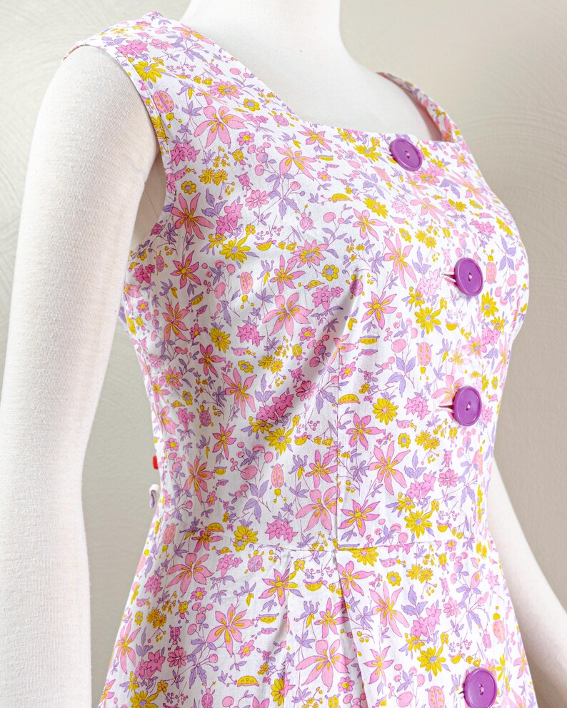 Puede incluir: Vestido sin mangas con fondo blanco y estampado floral en rosa, amarillo y morado. El vestido presenta un escote cuadrado, grandes botones morados y un corpi&ntilde;o entallado. El vestido se exhibe en un maniqu&iacute;.