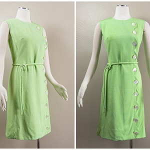 May include: A sleeveless green dress with a white button detail down the side. The dress has a tie belt and a fitted silhouette.