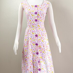 Puede incluir: Vestido de estilo vintage con estampado floral en tonos rosa, amarillo y morado sobre fondo blanco. El vestido presenta un escote cuadrado, mangas cortas y una botonadura frontal con grandes botones morados. Se muestra en un maniqu&iacute;.
