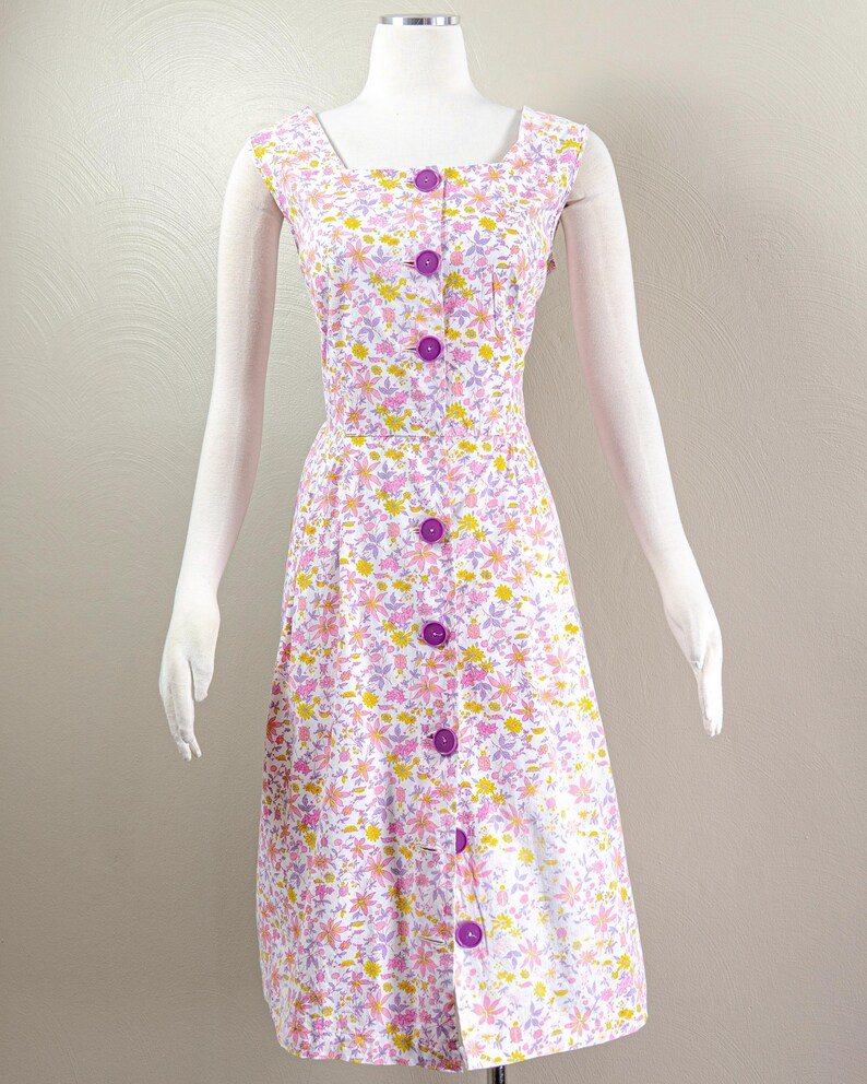 Puede incluir: Vestido de estilo vintage con estampado floral en tonos rosa, amarillo y morado sobre fondo blanco. El vestido presenta un escote cuadrado, mangas cortas y una botonadura frontal con grandes botones morados. Se muestra en un maniqu&iacute;.