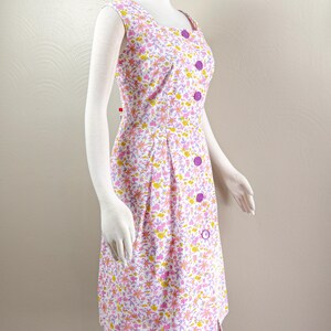 Puede incluir: Vestido sin mangas con estampado floral, escote cuadrado y botones morados. El vestido presenta un fondo blanco con un estampado de flores rosas, amarillas y moradas. El vestido se exhibe en un maniqu&iacute;.