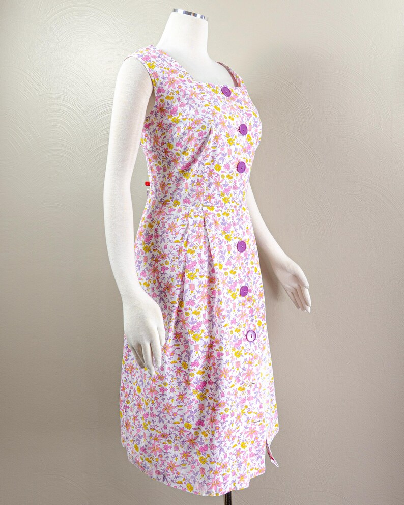 Puede incluir: Vestido sin mangas con estampado floral, escote cuadrado y botones morados. El vestido presenta un fondo blanco con un estampado de flores rosas, amarillas y moradas. El vestido se exhibe en un maniqu&iacute;.