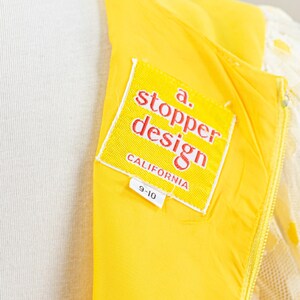 May include: Close-up of a yellow dress with a white lace overlay. The dress features a label that reads "a. stopper design CALIFORNIA" and a size tag of "9-10". The dress is on a mannequin.