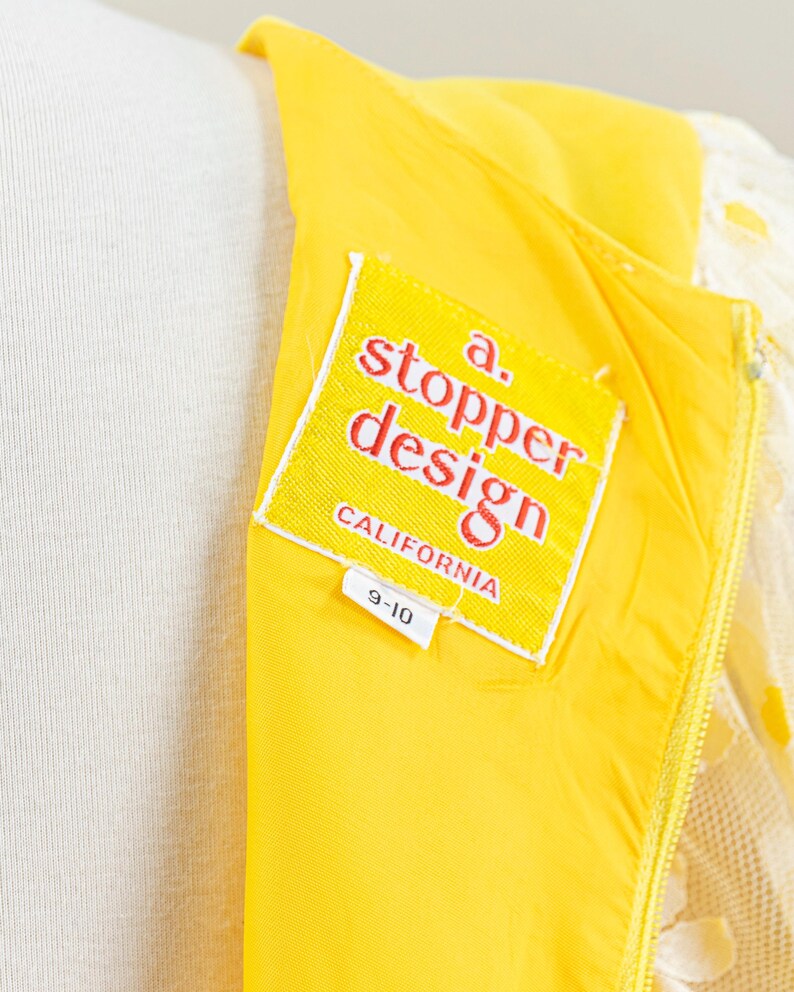 May include: Close-up of a yellow dress with a white lace overlay. The dress features a label that reads "a. stopper design CALIFORNIA" and a size tag of "9-10". The dress is on a mannequin.