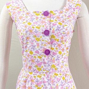 Puede incluir: Vestido sin mangas con cuello cuadrado y estampado floral en rosa, amarillo y morado sobre fondo blanco. El vestido tiene una botonadura frontal con botones morados grandes.