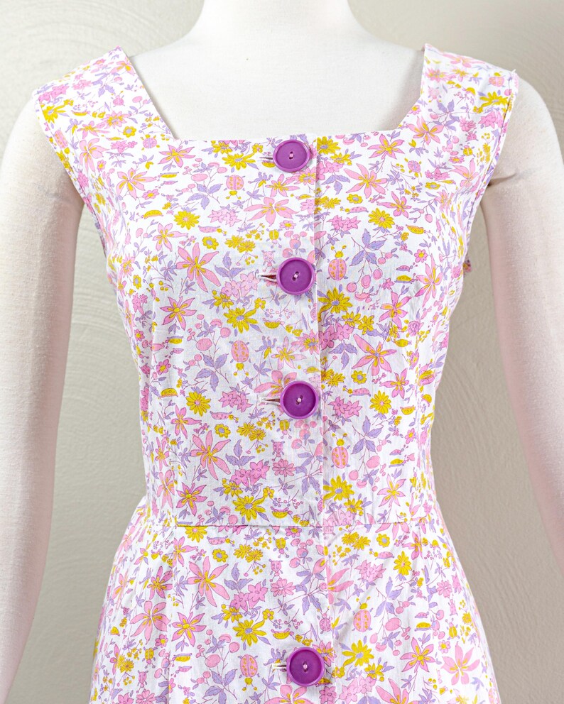 Puede incluir: Vestido sin mangas con cuello cuadrado y estampado floral en rosa, amarillo y morado sobre fondo blanco. El vestido tiene una botonadura frontal con botones morados grandes.