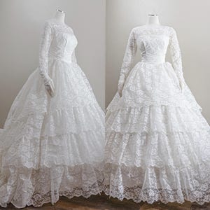 Puede incluir: Dos vestidos de novia de encaje blanco sobre maniquíes. Los vestidos presentan mangas largas, corpiños ajustados y faldas escalonadas con ribete de encaje. Un vestido tiene una larga cola. Los vestidos se exhiben sobre un fondo neutro.