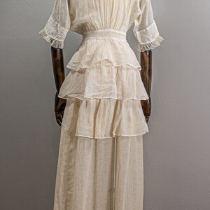 May include: A white cotton dress with a tiered skirt and a fitted bodice. The dress has a V-neckline and short sleeves with ruffles.