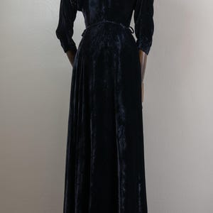 K&ouml;nnte beinhalten: Ein schwarzes Samt-Maxikleid mit Wickel-Oberteil und langen &Auml;rmeln. Das Kleid hat einen flie&szlig;enden Rock und einen hohen Ausschnitt.