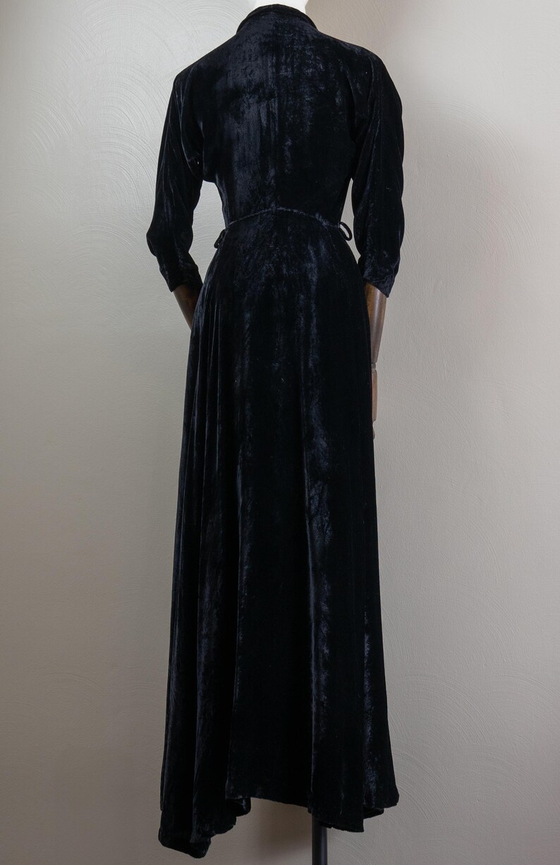 K&ouml;nnte beinhalten: Ein schwarzes Samt-Maxikleid mit Wickel-Oberteil und langen &Auml;rmeln. Das Kleid hat einen flie&szlig;enden Rock und einen hohen Ausschnitt.