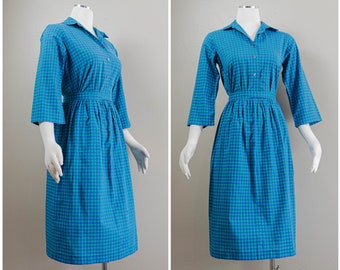 Könnte beinhalten: Ein blaues und grünes Gingham-Kleid mit Kragen, Dreiviertelärmeln und einer taillierten Taille. Das Kleid hat einen Knopfleisten vorne und einen Rock, der bis zum Knie reicht.